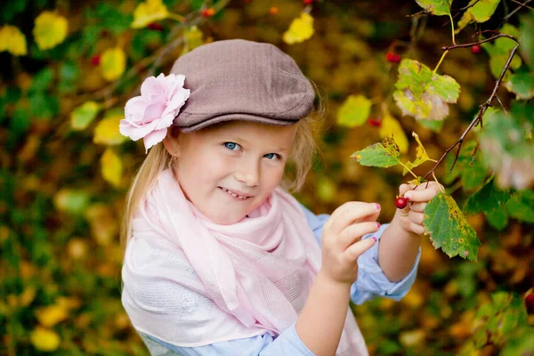 Una pequeña chica de ojos azules, de pelo claro, con gorra y bufanda rosa está arrancando un Rowan rojo de una rama. Mira la cámara, sonríe sonriente —  Fotos de Stock