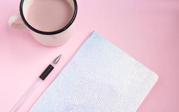 Holographic notebook and iron white cup with cocoa with pen on pink background. Flat lay. Selective focus