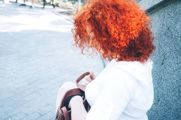 Ένα κορίτσι με λαμπερά σγουρά κόκκινα μαλλιά στο δρόμο κοιτάζει στην τσάντα της. — Φωτογραφία Αρχείου