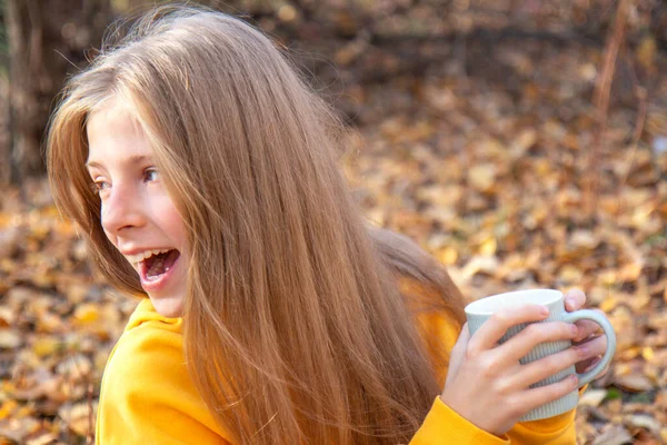 Krásné teen blondýny dívka v jasně oranžové mikiny s úsměvem šálek čaje v podzimním parku — Stock fotografie