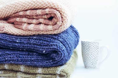 Bir yığın mavi, kahverengi, yeşil örgü süveter ve beyaz arka planda seramik beyaz bardak. Ev rahatlığı. Günlük kıyafet. Sonbahar havası. Sonbahar düzenli hafta sonu kavramı. Kopyalama alanı.