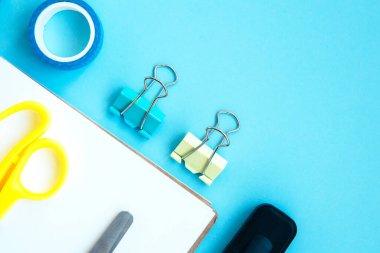 İşaretli beyaz defter, iki klemp, makas, klipsler ve mavi bir arkaplanda metin için yer olan viski. Okula hazırlanıyorum. Kırtasiyeli yaratıcı çalışma alanı. Okula dönelim. Düz Yatay