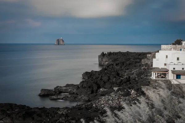 Ein Leuchtturm auf einer kleinen Insel, von einer Klippe mit schwarzen Felsen und einem weißen Gebäude aus gesehen. — Stockfoto