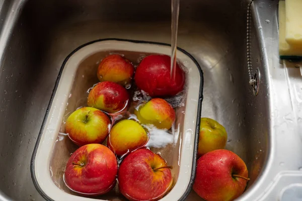 Apples in an enamel bowl under the flow water from tap in the sink