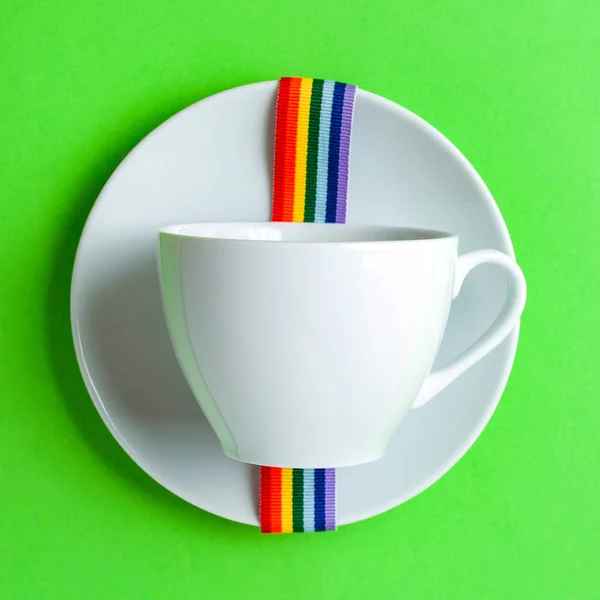 White coffee Cup on green pastel colorful paper geometric flat background with rainbow ribbon lgbt on saucer. Drink Cup template for your design to place text, image and logo layout