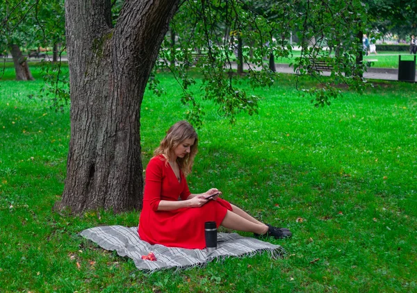 A young woman in a red dress is sitting under a tree in the Park. at the picnic thermos with coffee, apples uses smartphone