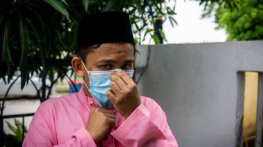 Coronavirus 'ta düzenlenen Bayram kutlamaları sırasında Baju Melayu kumaşı ve Songkok' ta 3 katmanlı maske takan Asyalı genç bir adamın portresi. Hijyen yaşam tarzı. Yeni normal kavram.