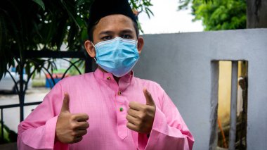 Coronavirus 'ta düzenlenen Bayram kutlamaları sırasında Baju Melayu kumaşı ve Songkok' ta 3 katmanlı maske takan Asyalı genç bir adamın portresi. Hijyen yaşam tarzı. Yeni normal kavram.