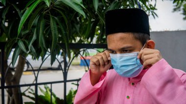 Coronavirus 'ta düzenlenen Bayram kutlamaları sırasında Baju Melayu kumaşı ve Songkok' ta 3 katmanlı maske takan Asyalı genç bir adamın portresi. Hijyen yaşam tarzı. Yeni normal kavram.