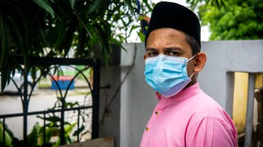 Coronavirus 'ta düzenlenen Bayram kutlamaları sırasında Baju Melayu kumaşı ve Songkok' ta 3 katmanlı maske takan Asyalı genç bir adamın portresi. Hijyen yaşam tarzı. Yeni normal kavram.
