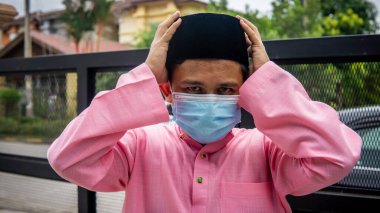 Coronavirus 'ta düzenlenen Bayram kutlamaları sırasında Baju Melayu kumaşı ve Songkok' ta 3 katmanlı maske takan Asyalı genç bir adamın portresi. Hijyen yaşam tarzı. Yeni normal kavram.