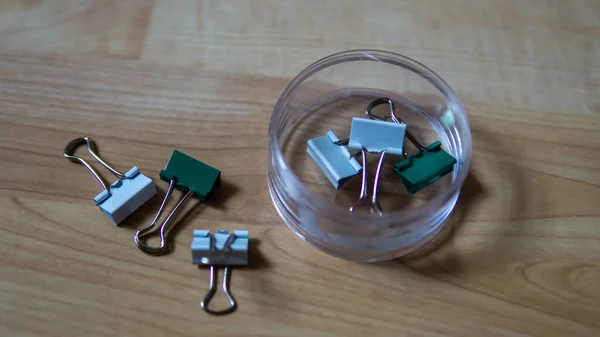 Top view of full binder clips in a transparent box and scattered on wooden background. It is a simple device for binding sheets of paper together. Selective focus on foreground.