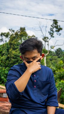 Geleneksel Baju Melayu kumaşlı genç bir Malezyalı 'nın portresi. Yeşil doğa arka planına sahip siyah kumaş bir maske takıyor. Virüs ve hastalıkların yayılmasını önlemek için koruma adımı.