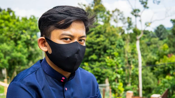 A portrait of a young Malay man with traditional Baju Melayu cloth wearing a black fabric face mask with green nature background. Protection step to avoid spreading viruses and diseases.