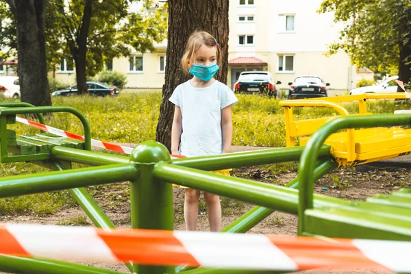 Маленькая милая девочка в маске на закрытой детской площадке. COVID-19 меры безопасности и защиты в городе . — стоковое фото