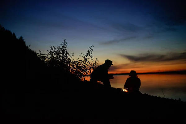 Die Menschen sitzen am Feuer. Picknick am Fluss. Sonnenuntergang — Stockfoto
