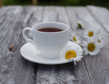 Tahta bir masada beyaz bir fincan çay. Masadaki papatyalar