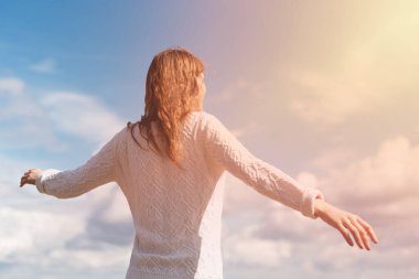 Beyaz kazak giyen kızıl saçlı bir kadın mavi gökyüzüne uzanmış kollarını açıyor..