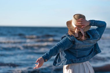 Denizin arka planında hasır şapkalı ve kot ceketli bir kadın. Arka planı kapat
