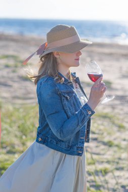 Hasır şapkalı kadın denizin kenarında dikiz aynasından bir bardak şarapla dikiliyor.