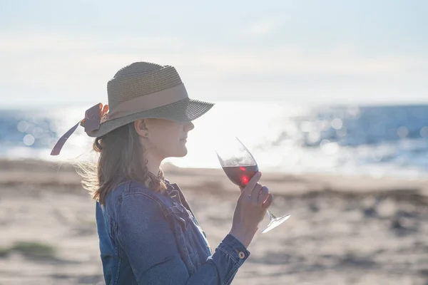 麦わらの帽子を被った女性がワインを片手に海のそばに立ち — ストック写真