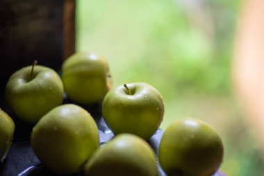 Yağmurdan sonra yağmur damlalarında yeşil elmalar siyah metal bir tahtaya saçılırlar ve arka planda yeşil çimenlere karşı hasır bir sepet vardır.