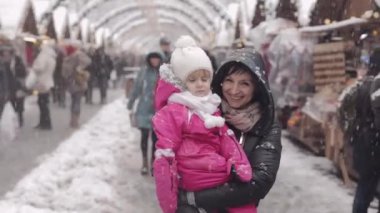 Anne ve kızı boyunca karlı havalarda sokakta yürüyor. Gülmek ve eğlenmek. Noel pazarı Fuarı. Komik küçük kız ve annesi.