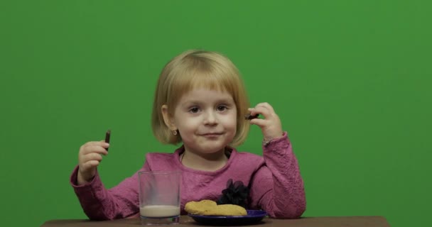 女孩坐在桌旁吃巧克力和饼干 快乐的三岁女孩 可爱的女孩微笑 漂亮的小孩3 4岁的金发女孩做鬼脸 绿色屏幕视频 色度键 — 图库视频影像