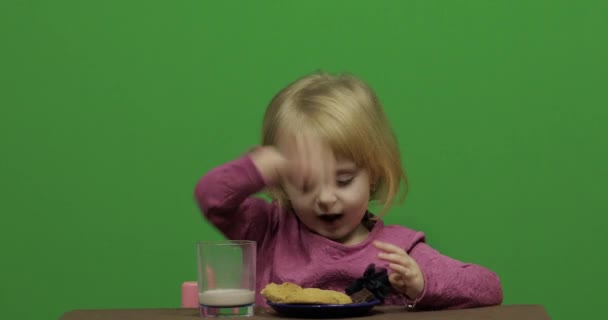 女孩坐在桌旁吃巧克力和饼干 快乐的三岁女孩 可爱的女孩微笑 漂亮的小孩3 4岁的金发女孩做鬼脸 绿色屏幕视频 色度键 — 图库视频影像