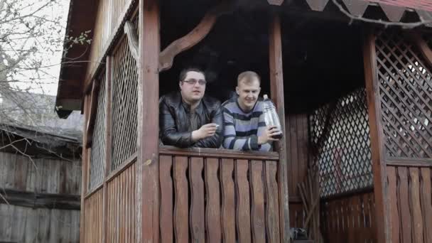 Двое мужчин на террасе пьют пиво из пластиковой бутылки и курят — стоковое видео