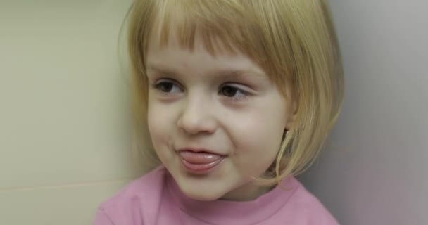 Primer plano de una pequeña rubia linda cara de chica. Chica sonriendo. Adentro. Retrato plano — Vídeos de Stock