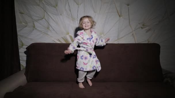 금발 머리와 귀여운 소녀는 소파에서 점프. 가운 의류입니다. 행복 — 비디오