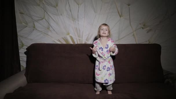 금발 머리와 귀여운 소녀는 소파에서 점프. 가운 의류입니다. 행복 — 비디오