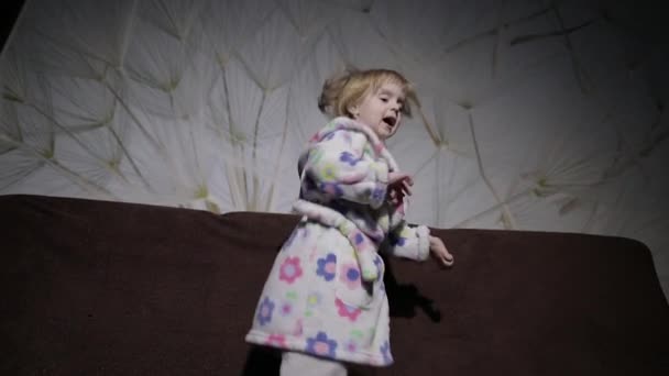 Маленькая милая девочка с светлыми волосами прыгает на диване. Одежда для халата. Счастливый — стоковое видео