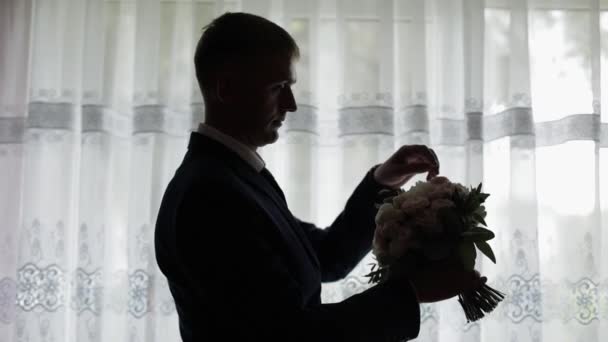 El novio guapo arregla su ramo de bodas cerca de la ventana. Silueta — Vídeos de Stock