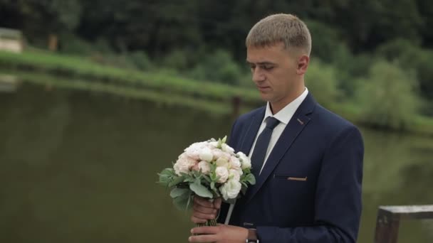 Novio con ramo de bodas en las manos en el parque. Camisa blanca, corbata negra, chaqueta — Vídeo de stock