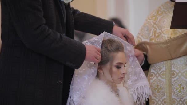 Κομψή νύφη σε μια παλιά εκκλησία. Ένας άντρας φοράει ένα κασκόλ στο κεφάλι του στη γυναίκα του. — Αρχείο Βίντεο