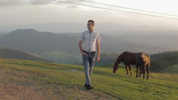 Młody człowiek stoi na polu niedaleko gór. Za nim pasą się konie. — Wideo stockowe