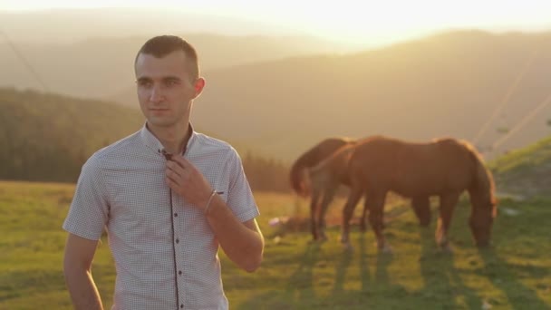 El joven está parado en el campo cerca de las montañas. Detrás de él pastando caballos — Vídeo de stock