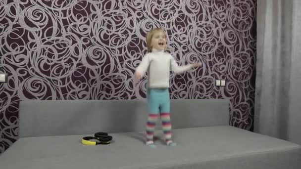 Little happy child jumping and dancing on sofa and listening music — Stock Video