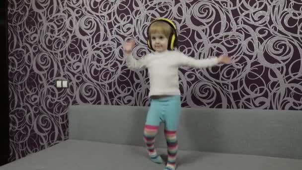Μικρό ευτυχισμένο παιδί άλμα και χορό στον καναπέ και ακούγοντας μουσική στα ακουστικά — Αρχείο Βίντεο