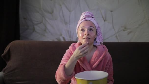 Egy nő késő esti filmet néz a TV-ben, popcornt eszik. Fürdőköpeny, arcpakolás — Stock videók