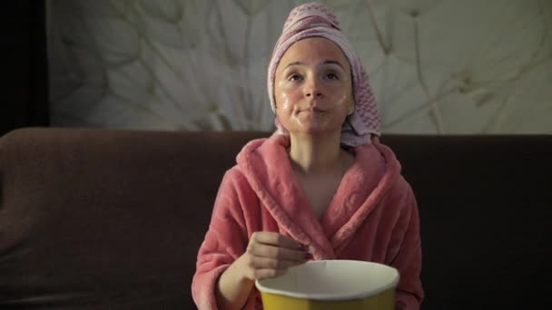 Mujer viendo una película nocturna en la televisión, comiendo palomitas de maíz. Albornoz, máscara facial — Vídeos de Stock