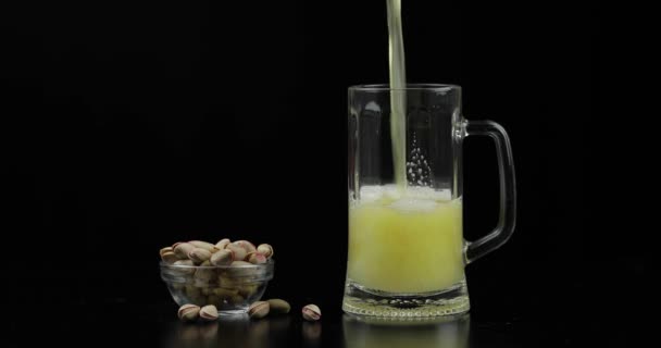 Bier ergießt sich in Glas auf schwarzem Hintergrund. Schüssel mit Pistazien-Nüssen — Stockvideo