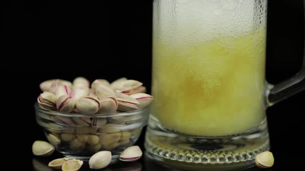 Beer is pouring into glass on black background. Bowl of pistachios nuts — Stock Video