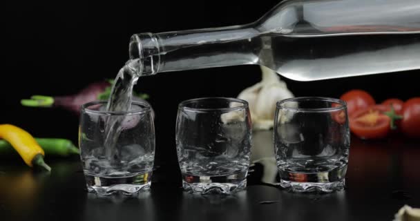 Verser la vodka alcoolisée d'une bouteille dans un verre à trois coups. Contexte avec des légumes — Video