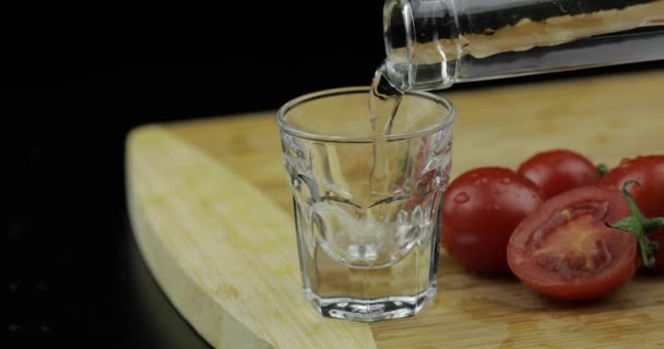 Вылейте водку из бутылки в рюмку на разделочную доску с помидорами — стоковое видео
