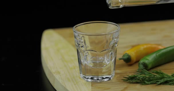 Wlać alkohol wódka z butelki w kieliszek na deski do krojenia z gorącym pieprzem — Wideo stockowe