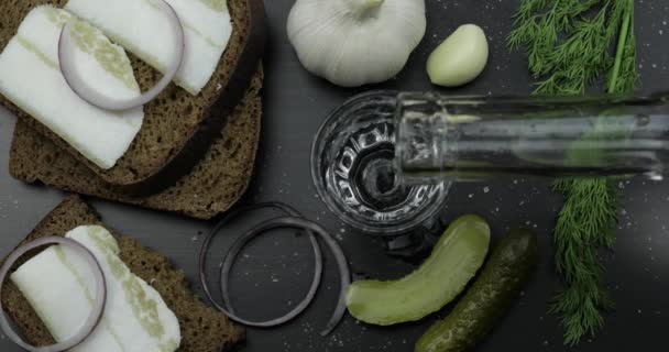 Vierta la bebida alcohólica vodka de una botella en un vaso de chupito. Superficie con snacks — Vídeos de Stock