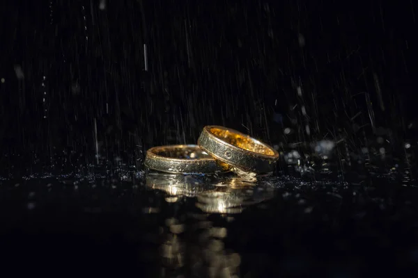 Wedding rings lying on dark surface shining with light. Water splashes — Stock Photo, Image